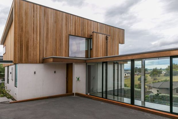 Exterior view of a contemporary two-storey house with vertical timber cladding, rendered lower walls, projecting upper-level form, and extensive floor-to-ceiling glazing. The paved driveway or terrace occupies the foreground, with a suburban landscape visible beyond the glass.