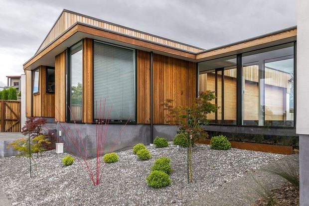 Contemporary single-storey house exterior with warm vertical timber cladding, dark-framed floor-to-ceiling glazing, a low-pitched irregular roofline, and a minimalist gravel garden with sculpted shrubs. The home is photographed from the front courtyard under an overcast sky.