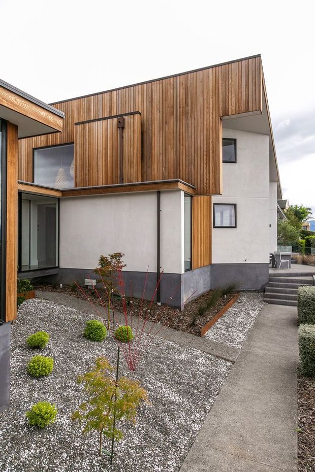 Exterior view of a contemporary multi-level house with a sharply angled roofline, extensive vertical timber cladding, white rendered walls, dark base trim, and large glazed openings. A minimalist landscaped courtyard uses gravel beds, clipped shrubs, young trees, and concrete paths with steps leading around the building.