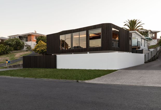 Street-level exterior view of a contemporary single-storey home with a dark vertically clad upper facade, large glazed openings, a white rendered boundary wall, and a sloping concrete driveway. The house sits within an established suburban setting with neighbouring homes and palm planting visible around it.