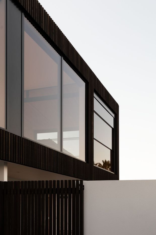 Close-up exterior view of a contemporary two-storey building with expansive dark-framed glazing, dark vertical slatted cladding and a projecting upper volume. A white boundary wall and dark slatted gate occupy the foreground, while warm interior ceiling surfaces and reflected vegetation are visible through the windows.