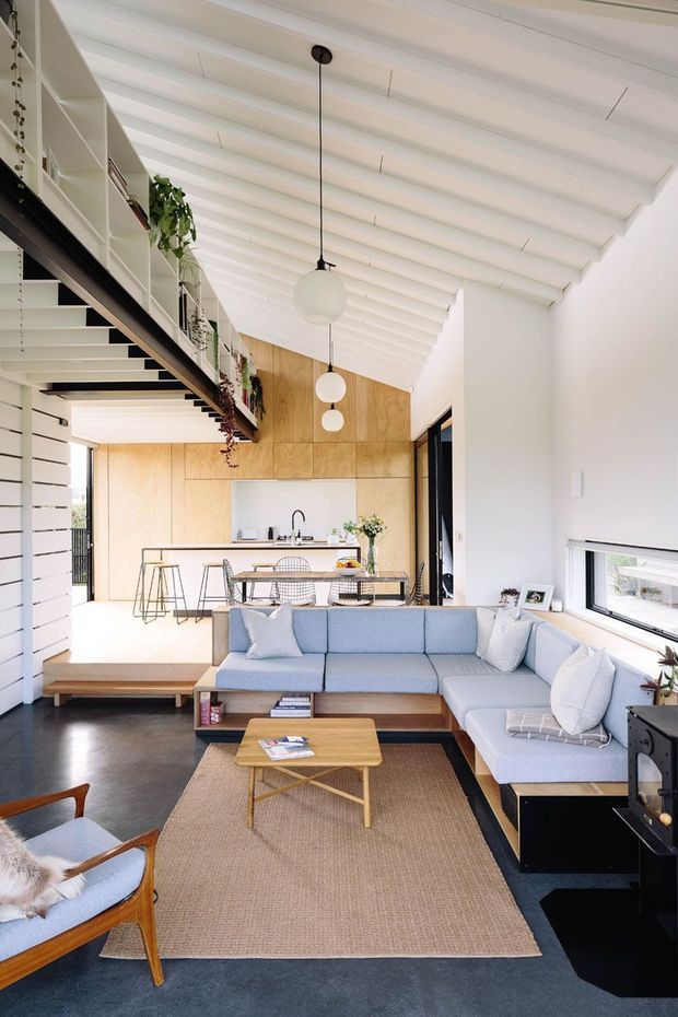 A bright open-plan living, dining and kitchen interior with a double-height sloping ceiling and exposed white-painted rafters. The room features a large pale blue L-shaped sectional sofa, timber coffee table, woven rug, timber-framed lounge chair, compact dining table with wire-frame chairs, and a white kitchen with a timber island. Warm timber wall panelling forms the kitchen backdrop, while a mezzanine-style shelving and walkway with trailing plants runs along the left side. Globe pendant lights hang in a row, and large windows provide natural light.