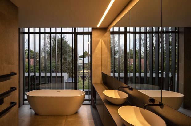 A luxurious contemporary bathroom with a freestanding oval bathtub positioned in front of floor-to-ceiling black-framed glazing. A long dark vanity with integrated basins runs along the right wall, which is clad in warm brown stone or porcelain panels and lined with a large mirror. Linear recessed lighting creates a warm, dramatic atmosphere, while trees and an outdoor area are visible beyond the glazing.