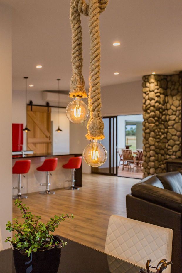 Warm open-plan residential interior featuring a kitchen and living area. Two prominent rope-hung pendant lights with exposed glowing bulbs hang in the foreground. The kitchen has a dark island with bright red bar stools, timber flooring, a timber barn-style sliding door, and recessed ceiling lighting. A rounded natural-stone feature wall and sliding glass doors lead to an outdoor dining area. A dark leather sofa and quilted dining chair are visible in the foreground.