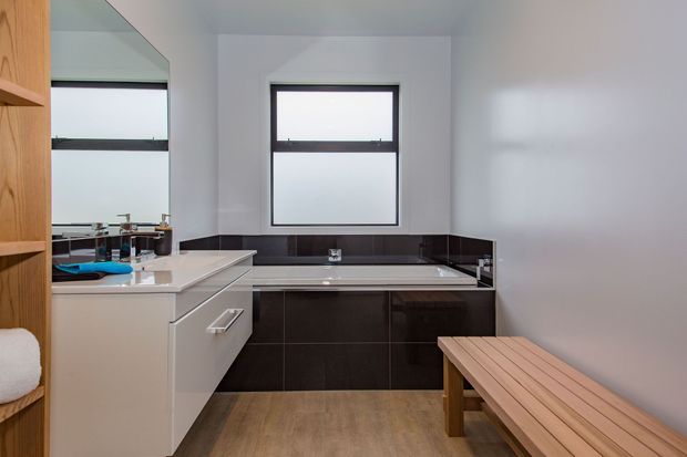 A compact contemporary bathroom with a freestanding bathtub beneath a black-framed window, dark tiled bath surround and vanity cabinetry, white walls, and warm timber-look flooring. A timber bench occupies the foreground, while a large mirror reflects the vanity and basin area.