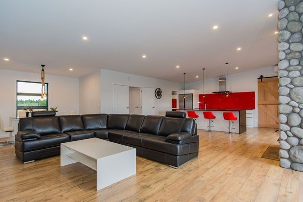 Open-plan contemporary living and kitchen area with a large black leather sectional sofa, white rectangular coffee table, warm timber flooring, recessed ceiling lighting, and a vivid red kitchen island and cabinetry with red bar stools. A substantial rounded-stone feature column and timber barn-style door add rustic natural accents.