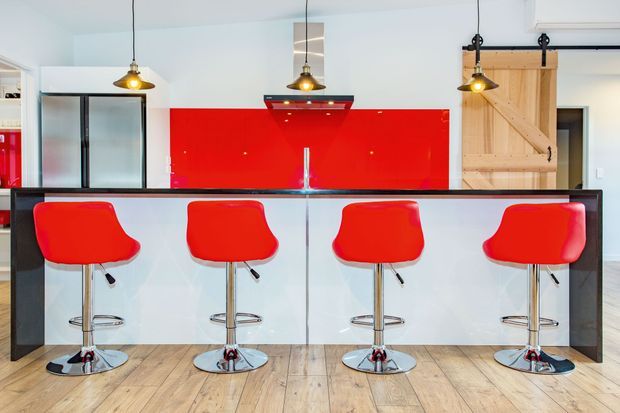 Bright contemporary kitchen featuring a long island with a dark countertop, white cabinetry or panelled frontage, and four vivid red bar stools. A glossy red glass splashback, stainless-steel rangehood, pendant lighting, timber flooring, and a sliding barn-style timber door create strong industrial-modern accents.