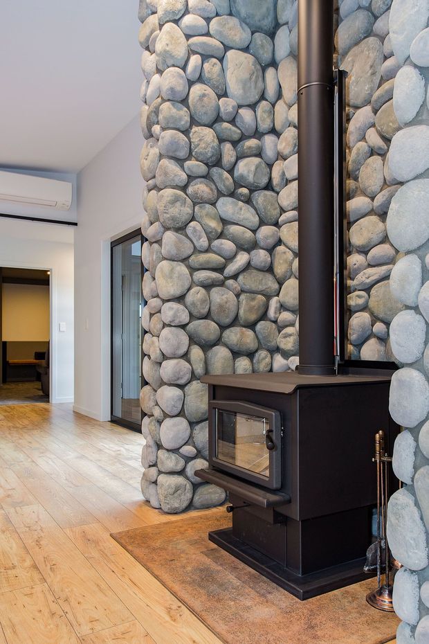Interior view of a rustic living area featuring a freestanding black wood-burning stove with flue, set against a prominent rounded natural river-stone feature wall. Pale timber flooring, white walls, glazing, and an adjoining room are visible.
