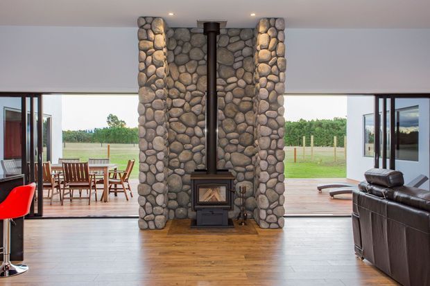 Open-plan living room with a central freestanding wood-burning fireplace and tall rounded river-stone masonry surround. Dark timber flooring, broad glazed openings and folding doors connect the room to a timber deck and rural landscape. Dining furniture is visible to the left and leather seating to the right.