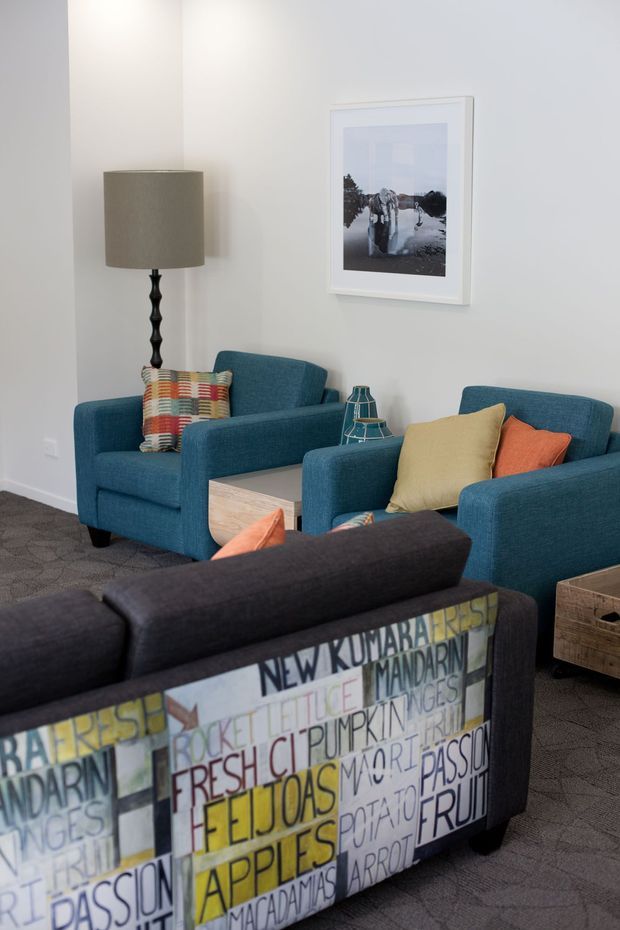 A bright, contemporary sitting room with two teal upholstered armchairs and a matching sofa arranged around a small light-wood table. The room has pale walls, dark patterned carpet, a tall floor lamp, colourful cushions, a decorative wire lantern, framed artwork, and a distinctive typographic upholstered sofa back in the foreground.