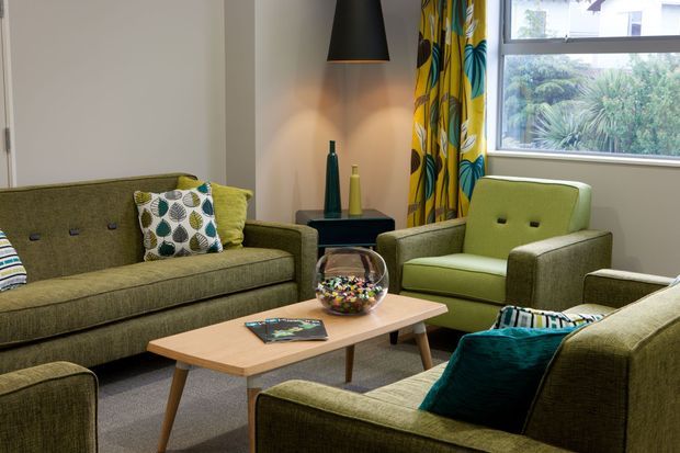 A cosy living room furnished with olive-green upholstered sofas and armchairs arranged around a light timber coffee table. Patterned cushions, a teal accent cushion, a dark side table, sculptural vases, a black pendant light, and bold yellow-and-green botanical curtains add colour and character. A large window provides natural light, with a neutral carpet and pale walls completing the space.