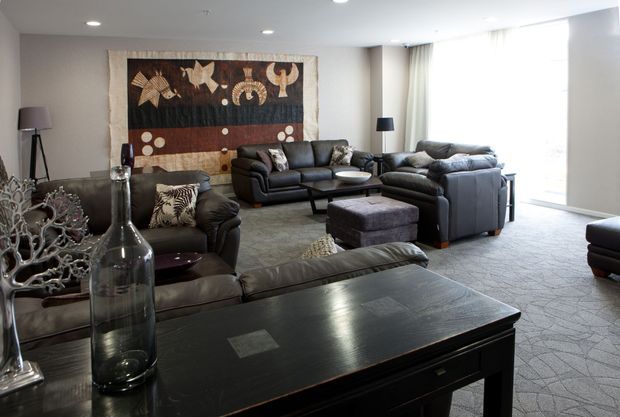 Spacious contemporary living room furnished with multiple dark leather sofas and armchairs arranged around low tables and ottomans. A large decorative wall textile provides a feature backdrop, while full-height sheer curtains and recessed ceiling lights create a bright, polished interior. A dark timber console table with glass and metallic decorative objects occupies the foreground.