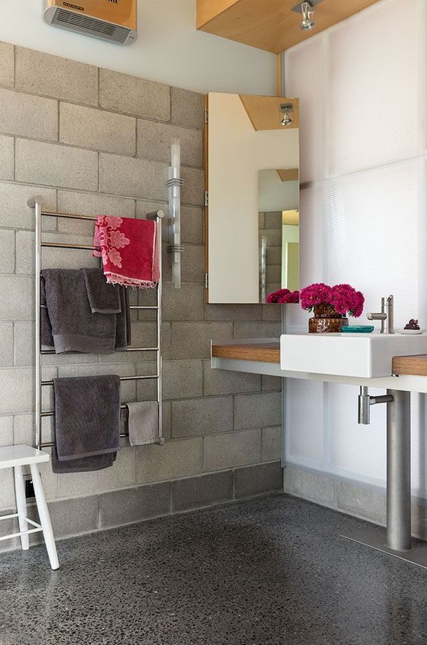 Contemporary bathroom with exposed grey concrete block walls, a dark polished aggregate floor, floating timber-edged vanity with a white rectangular basin, wall-mounted chrome tapware, mirrored cabinet, towel ladder, and translucent window blinds. Bright pink flowers provide a vivid accent against the industrial material palette.