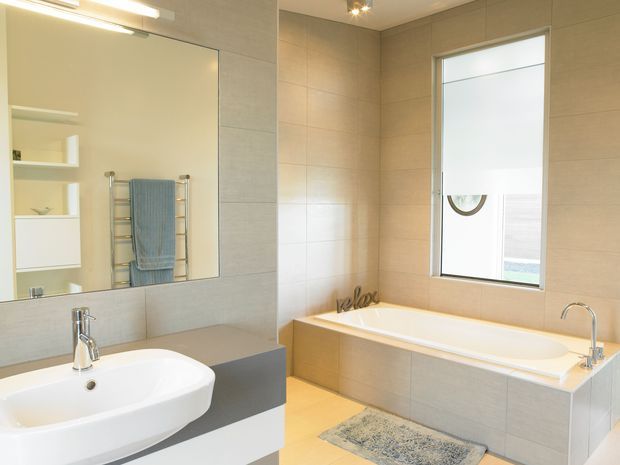 Bright contemporary bathroom with a freestanding-style built-in bathtub beneath a large frosted window, a white vessel basin on a grey vanity, wall-mounted shelving, a chrome towel rail, and pale beige tiled walls and floor. The space is clean, light-filled, and minimally styled.