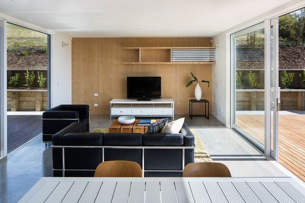 Bright, compact contemporary living room and dining area with black leather seating arranged around a low coffee table, a wall-mounted television, and a full-height timber feature wall with open shelving and a concealed storage unit. Large glazed folding or sliding doors open to timber decking and landscaped outdoor areas on both sides, bringing in abundant natural light. A pale concrete floor, white walls, and simple black and timber furnishings create a clean, modern indoor-outdoor setting.