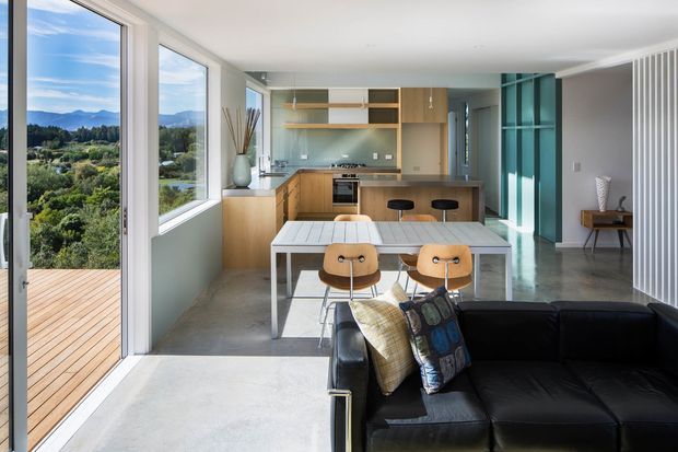 Bright open-plan interior combining a compact kitchen, dining area and living space. Full-height glazing opens to a timber deck and expansive green, mountainous views. The room features light timber cabinetry, a pale stone or concrete-look floor, a turquoise built-in shelving wall, a white dining table with timber chairs, and a black leather sofa. Vertical white wall detailing adds texture along the right side.