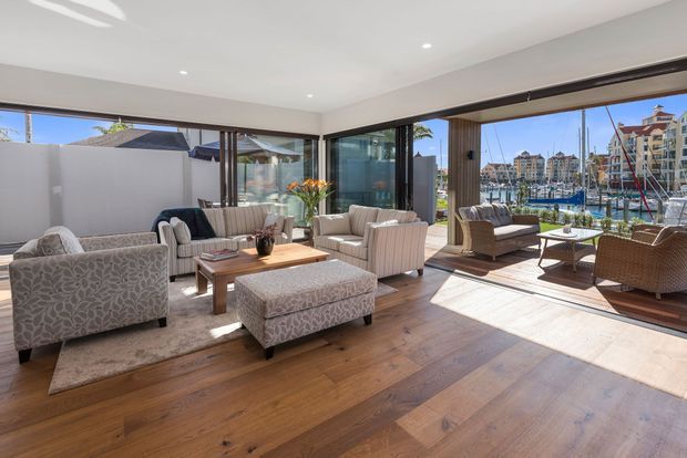 Bright, spacious waterfront living room with expansive corner glazing and folding glass doors opening to a marina view. The room features warm timber flooring, neutral upholstered sofas and armchairs, a patterned ottoman, a timber coffee table, recessed ceiling lights, and a relaxed contemporary coastal feel.