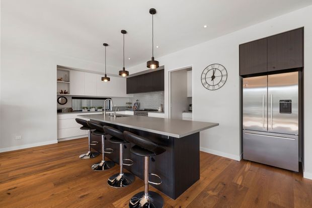 Bright contemporary kitchen with a large island and four black bar stools, white cabinetry, dark wood-toned cabinetry, stainless-steel appliances, pendant lighting, a tiled backsplash, and warm timber flooring. An open doorway leads to an adjacent room.