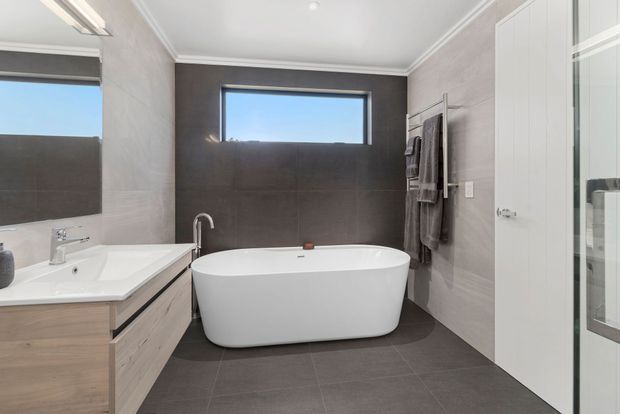 Contemporary bathroom featuring a freestanding white bathtub beneath a narrow horizontal window, a floating light-wood vanity with white basin, grey tiled walls and floor, towel rails, and a glass-enclosed shower at right. The room is bright, clean, and restrained in its material palette.