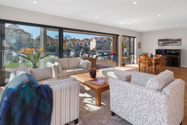 Bright, spacious living room with large sliding glass doors opening to a waterfront promenade or marina-facing outlook. The room features multiple upholstered sofas and armchairs, a timber coffee table, a timber dining table with chairs, polished timber flooring, recessed ceiling lights, and neutral patterned furnishings. A piano and framed artwork sit along the rear wall, while strong daylight fills the space.