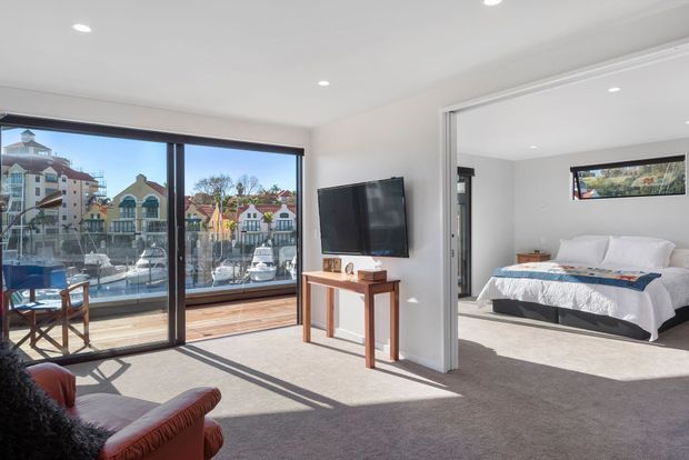 Bright open-plan living area with a large glazed balcony frontage overlooking a marina, connected directly to a carpeted bedroom. The living room has a wall-mounted television, slim timber console, leather lounge seating and timber decking at the glazing. The bedroom contains a low-profile bed with white bedding and a narrow horizontal window. The interior is simple and contemporary with neutral finishes and abundant daylight.