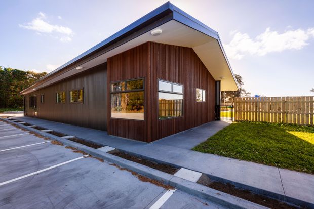 Single-storey contemporary building with a sharply pitched gable roof, dark vertical timber cladding, and large rectangular windows. The structure sits beside a paved parking area and concrete walkway, with a fenced grassy area to the side. The exterior has clean, functional lines and strong geometric detailing.