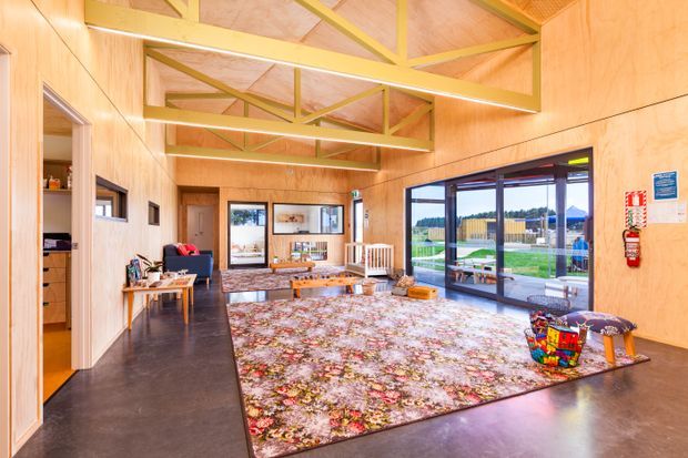Bright, open-plan communal living space with warm timber wall and ceiling lining, exposed yellow-painted roof trusses, polished dark concrete flooring, extensive black-framed glazing, and a large floral area rug. The room includes a lounge seating area, low coffee table, children's furniture and artwork, with views to a landscaped outdoor area through the glazing.