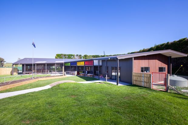 Exterior view of a low, single-storey contemporary building with a long horizontal form, shallow sloping roof, dark cladding, and colourful glazed sections. The building fronts a large manicured lawn with curved concrete paths, fencing, and a flagpole; a landscaped green roof or planted roof edge is visible along the rear roofline.