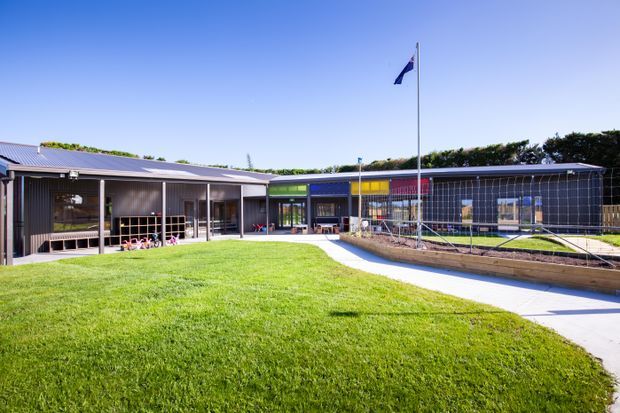 Wide exterior view of a low-rise contemporary institutional building arranged around a landscaped lawn and paved walkway. The complex has dark metal-clad façades, extensive glazed sections, colorful translucent window panels, covered outdoor verandas, and a fenced recreational or sports area. A flagpole stands prominently in the central courtyard-like lawn.