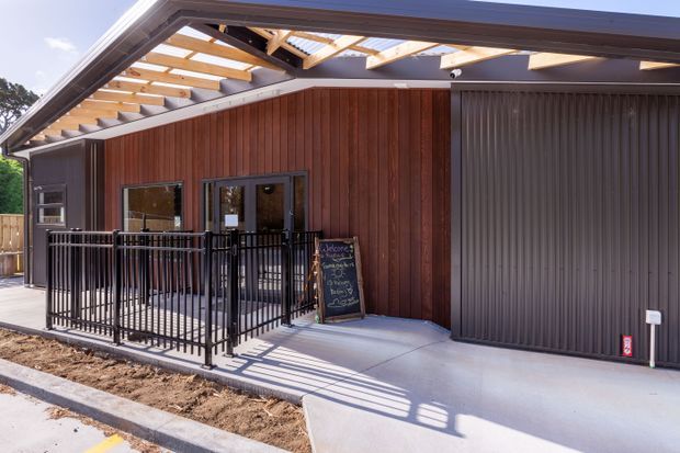 Exterior view of a contemporary single-storey commercial-style building with a covered entry terrace, black metal balustrade, vertical timber wall cladding, dark ribbed metal cladding, glazed entry doors, and a broad concrete forecourt. Exposed timber roof framing and translucent roof panels create a bright sheltered outdoor threshold.