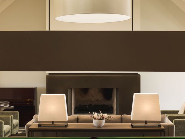 A refined living room centered on a large fireplace with a dark rectangular surround and substantial horizontal architectural beam or divider above. A long console table with two matching table lamps sits in front of the fireplace, with a sofa, upholstered armchairs, a piano, and a large suspended circular pendant light visible. The palette is warm and neutral with dark brown, cream, taupe, and olive accents.