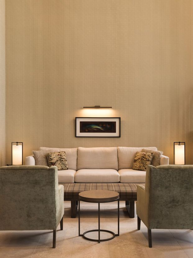 A refined living room arranged around a beige upholstered sofa and two plush armchairs, with a low round coffee table, patterned ottoman or bench, framed artwork, slim table lamps, and a tall warm wood-toned wall finish. The room has a warm, softly lit, symmetrical composition with tiled flooring and understated decorative styling.