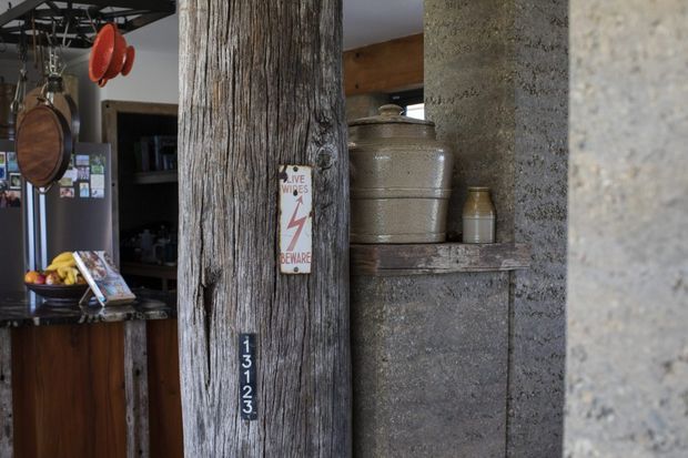 Close-up interior view of a rustic kitchen or living space featuring a heavily textured reclaimed timber post, rough concrete or masonry wall, dark timber cabinetry, and a stone or concrete ledge displaying ceramic vessels. Industrial hanging cookware and vintage signage add an eclectic, utilitarian character.