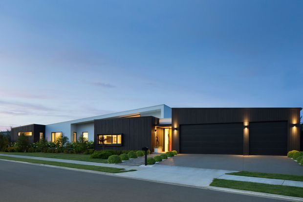 Contemporary single-storey residential facade photographed from across the street at dusk. The home has a strong horizontal composition, a pale rendered or concrete-like main volume, dark vertically ribbed cladding to the projecting entry and double garage, a low-pitched or flat roofline, warm illuminated windows and entry, manicured planting, and a broad concrete driveway.