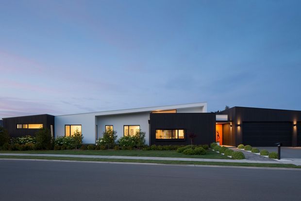 A low-profile contemporary single-storey residence viewed from across the street at dusk. The facade combines dark ribbed cladding with smooth white rendered volumes, warm illuminated windows, a recessed entry, landscaped planting, a stepped front path, and an integrated dark double garage. The flat-roof composition and horizontal arrangement create a clean modern suburban exterior.