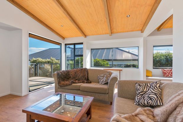 Bright living room with warm timber-lined exposed beam ceiling, white walls, timber flooring, large windows and glazed doors opening to a balcony or terrace. A brown sofa and armchairs surround a glass-topped timber coffee table, with patterned cushions and soft throws adding visual interest. The room overlooks neighbouring pitched roofs and greenery.