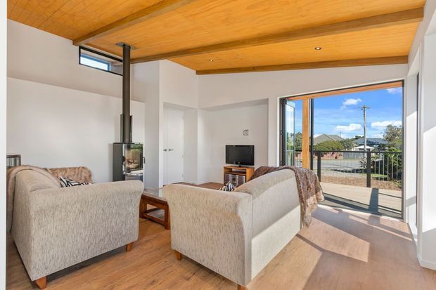 Bright contemporary living room with a compact wood-burning stove, timber flooring, a warm timber-lined ceiling with exposed beams, and pale upholstered seating arranged around a small television. Large glazed doors open to a balcony or terrace with suburban views, bringing in abundant natural light.