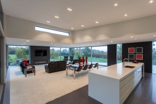 A spacious open-plan contemporary interior combining a white kitchen with a large island, dining area, and living room. Floor-to-ceiling glazing wraps around the room, opening views to a landscaped outdoor setting. The room features dark tiled flooring, light carpet, white cabinetry and benchtops, a dark feature media wall, recessed ceiling lights, and a high ceiling with clerestory glazing.