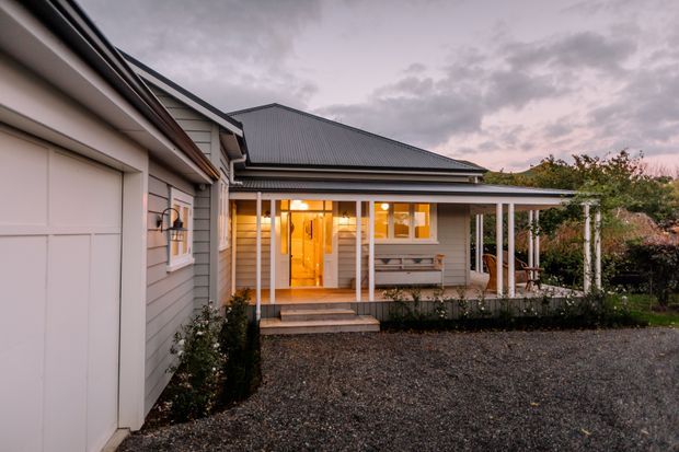 Exterior view of a single-storey weatherboard home at dusk, featuring a large garage door, pitched corrugated metal roof, covered front veranda with slender posts, timber steps, and warmly lit entry glazing. A gravel driveway and landscaped garden surround the house.