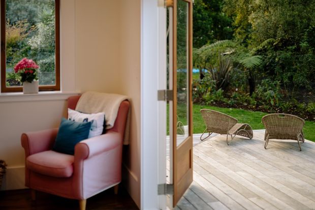 A cozy interior sitting area with a pink upholstered armchair, cushions, a small potted plant, and a timber-framed window. An open glazed timber door leads directly to a spacious pale timber deck with two woven lounge chairs, overlooking a lush landscaped garden.