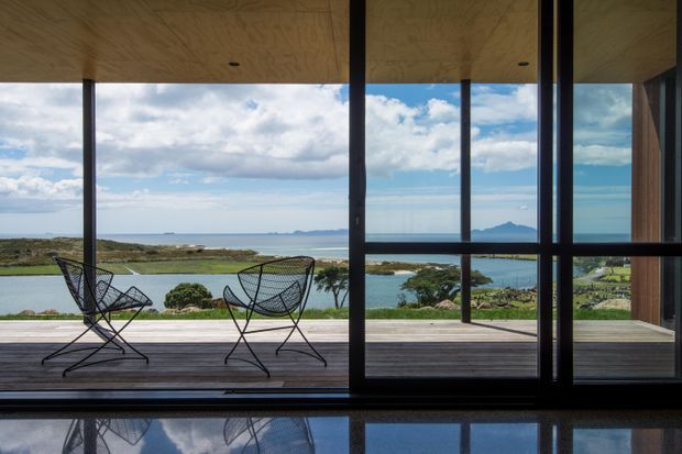 A minimalist contemporary interior opens onto a broad timber deck through expansive dark-framed glazing. Two wire-frame lounge chairs sit on the deck, while the view beyond includes coastal water, low vegetation, distant islands or headlands, and a partly cloudy sky. The interior features a warm timber-lined ceiling, dark structural framing, and a highly reflective dark floor.