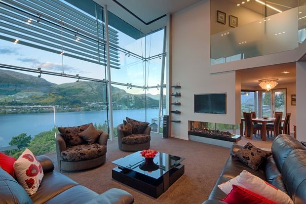 A sophisticated double-height living room with floor-to-ceiling glazing overlooking a lake and mountainous landscape. The room features leather sofas and patterned armchairs arranged around a glossy black coffee table, a built-in linear fireplace beneath a wall-mounted television, a dining area beyond, and an upper-level mezzanine or landing with glass balustrading. Horizontal external sun-shading fins are visible across the glazing.