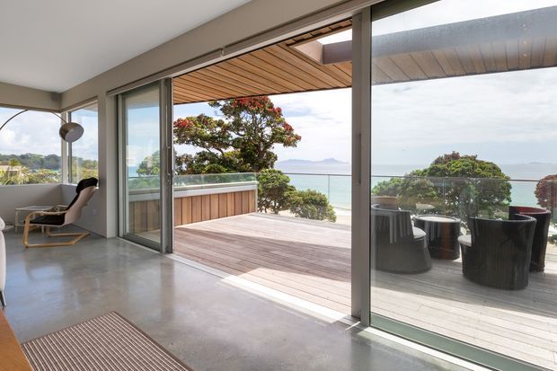A modern open-plan interior with polished concrete flooring and expansive sliding glass doors opening onto a timber deck. The deck includes outdoor seating and a covered timber-lined ceiling, with landscaped planting and a broad coastal or waterfront view beyond. A minimalist lounge chair and pendant light are visible inside.