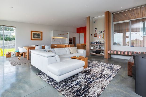 Open-plan contemporary living area with a large white sectional sofa, patterned area rug, dining table, and an integrated kitchen visible behind it. The space features polished concrete flooring, white walls, warm timber wall panelling and columns, extensive glazing, built-in shelving, and a timber-lined window seat. A red kitchen splashback and cabinetry provide a strong accent.