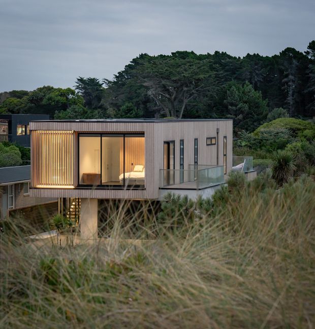 A contemporary elevated residential pavilion set within dense coastal vegetation, viewed from the surrounding landscape at dusk. The compact rectangular building has vertical timber cladding, a flat roof, extensive dark-framed glazing, a projecting glazed balcony, and a warmly lit bedroom visible through the windows. Native grasses and established trees surround the structure.