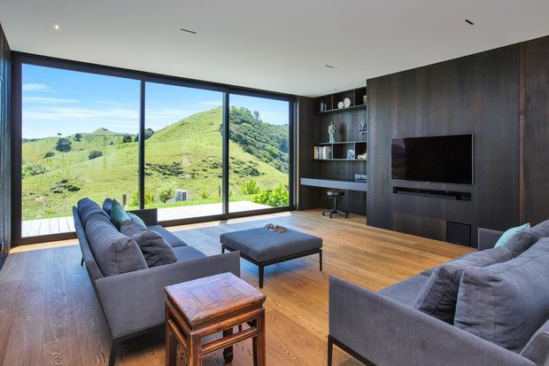 Contemporary living room with expansive floor-to-ceiling glazing overlooking rolling green hills. The room features dark timber wall panelling and built-in shelving with a television, warm timber flooring, two large grey sofas, a central upholstered coffee table, and a compact timber side table.
