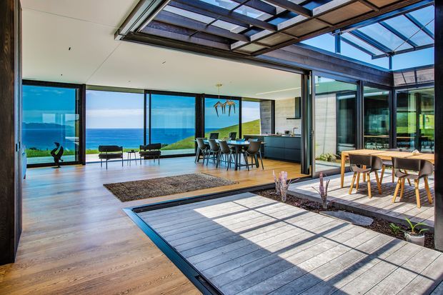 A bright, open-plan contemporary home with expansive floor-to-ceiling glazing overlooking the ocean. The interior combines a living area and dining kitchen with dark-framed glass walls that open onto a central timber-decked courtyard. A glazed overhead structure creates a covered indoor-outdoor transition, while timber flooring, dark cabinetry, modern dining furniture, and a neutral rug complete the space.