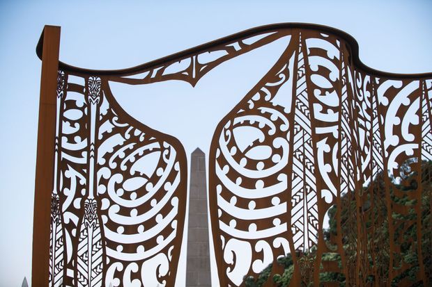 Close-up exterior view of an ornate rust-brown decorative metal gate or screen with flowing organic cut-out motifs and curved upper edges. A slender stone or concrete obelisk-like feature and trees are visible behind it against a pale blue sky.