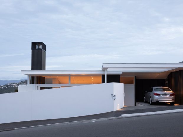 Street-facing view of a low-profile contemporary residence at dusk, featuring crisp white rendered walls, broad horizontal roof planes, extensive warm-lit glazing, a tall dark vertical chimney or tower element, and a partially enclosed dark carport with a parked car. The home sits on a sloping suburban site with distant hills visible beyond.