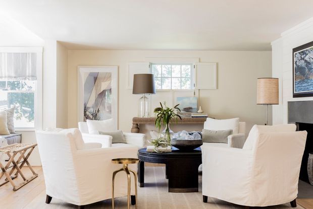 Bright, refined living room with slipcovered white lounge chairs and sofa arranged around a dark round coffee table. The space features warm neutral walls, pale timber flooring, a central console beneath a small multi-pane window, layered table and floor lamps, framed artwork, and a fireplace surround at right. Natural light creates an airy, comfortable atmosphere with restrained traditional styling.